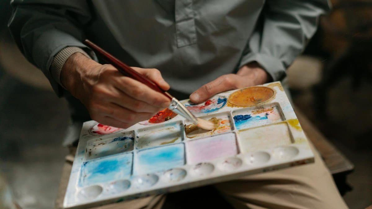Close-up of an artist's hands mixing colors on a watercolor palette with a paintbrush.