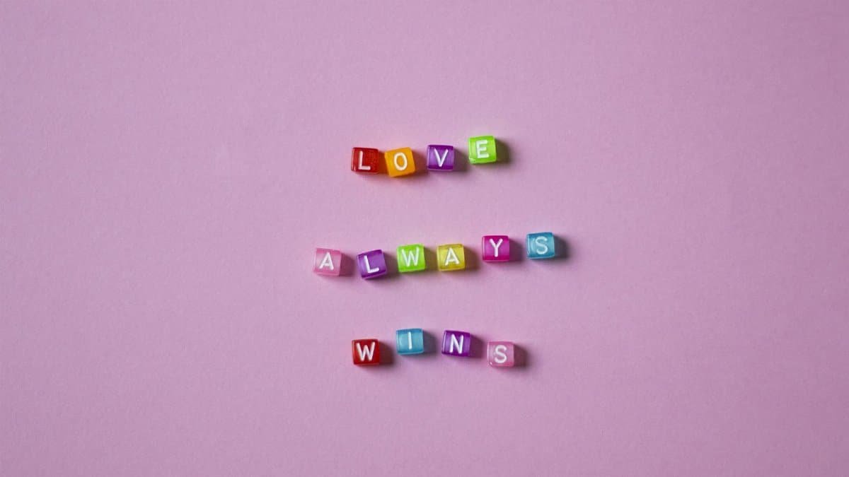 Vibrant colorful letter beads spelling 'Love Always Wins' on a soft pink background.