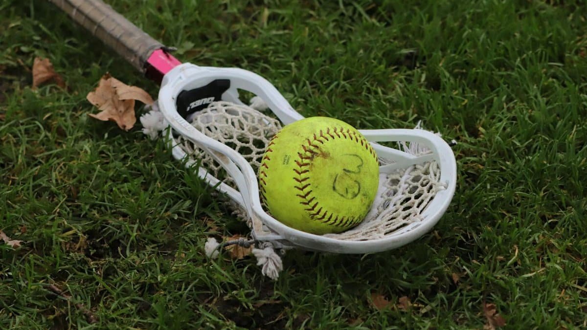 A softball nestled in a lacrosse stick lying on green grass, highlighting sports crossover.