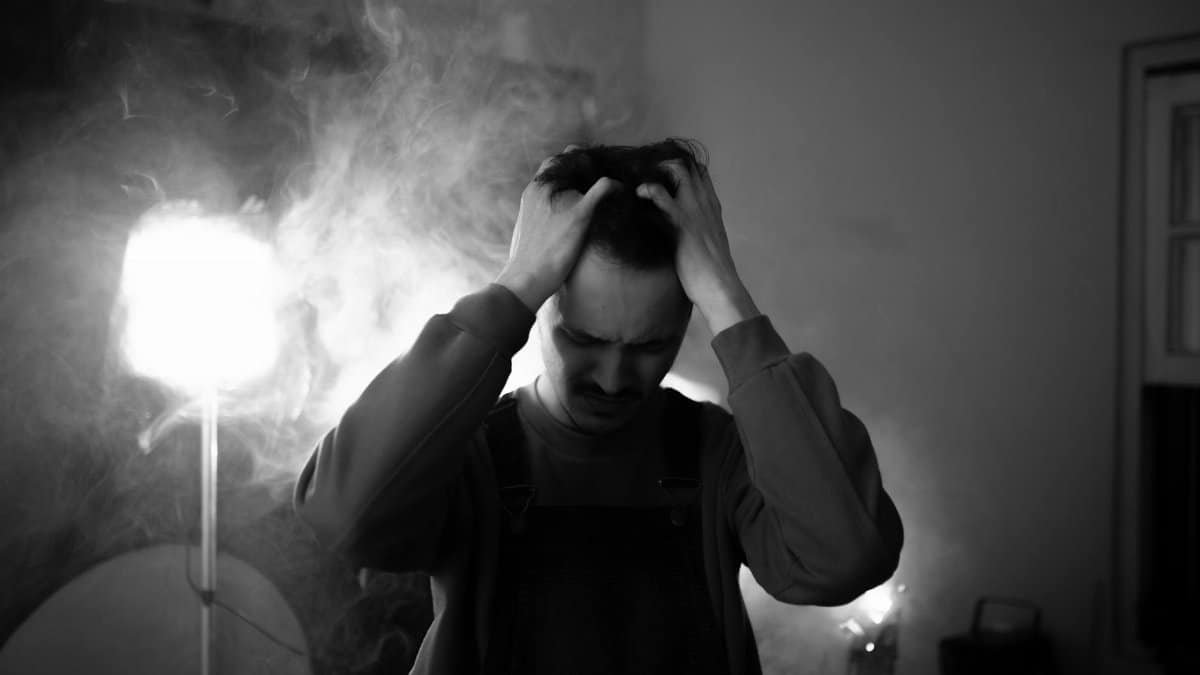 Black and white of upset male standing in room with lightning equipment and smoke while thinking about difficult decision at home