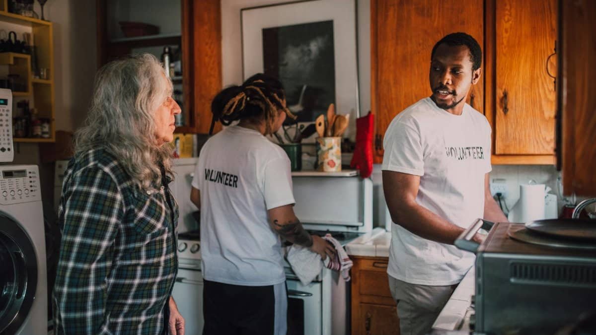 Volunteers interact and assist a senior in a cozy kitchen environment, promoting community support.
