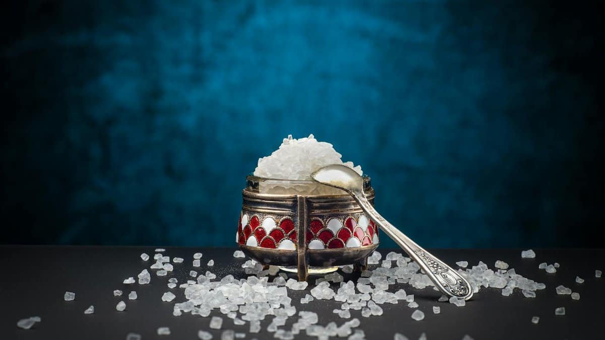 A close-up shot of rock salt in a decorative container with a silver spoon on a blue background.