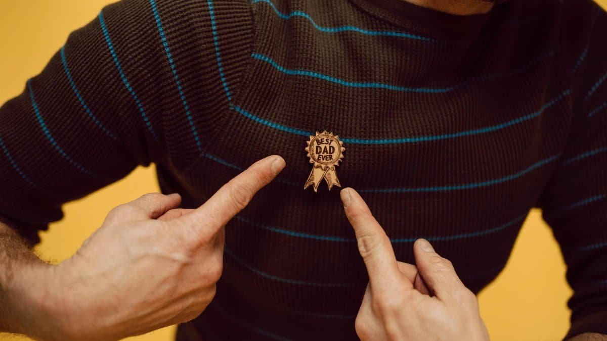 A man proudly highlighting his 'Best Dad Ever' badge on a striped sweater against a yellow background.