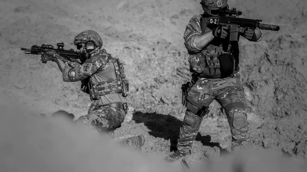 Two soldiers in camouflage and gear in a desert setting, engaged in a tactical action.