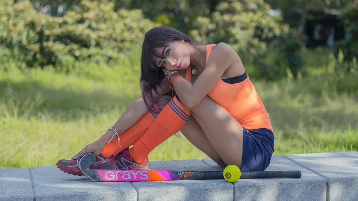 A young woman in orange sportswear sitting outdoors with field hockey equipment on concrete.