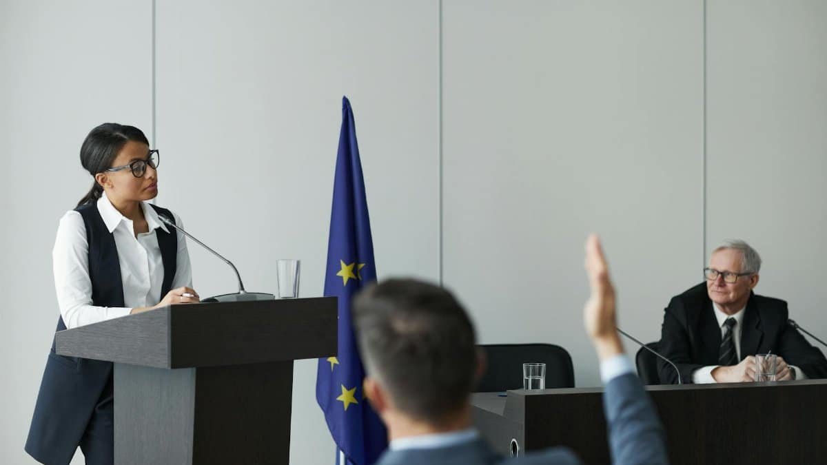 A woman speaking at a European Union conference while delegates listen attentively.