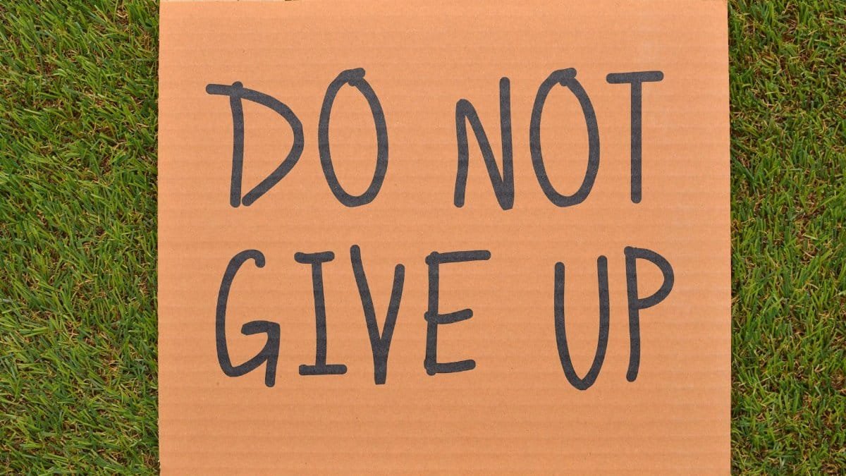 Cardboard sign reading 'Do Not Give Up' placed on green turf, symbolizing perseverance.
