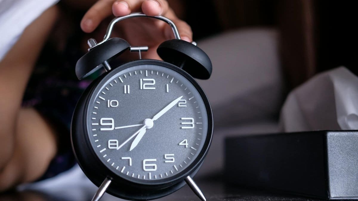 A hand reaches out to turn off an analogue alarm clock in the early morning.