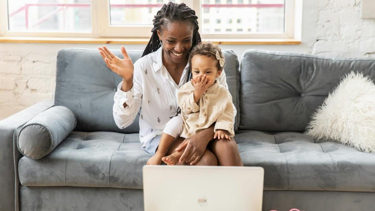 A joyful mother and daughter wave during a video call from their cozy living room couch.