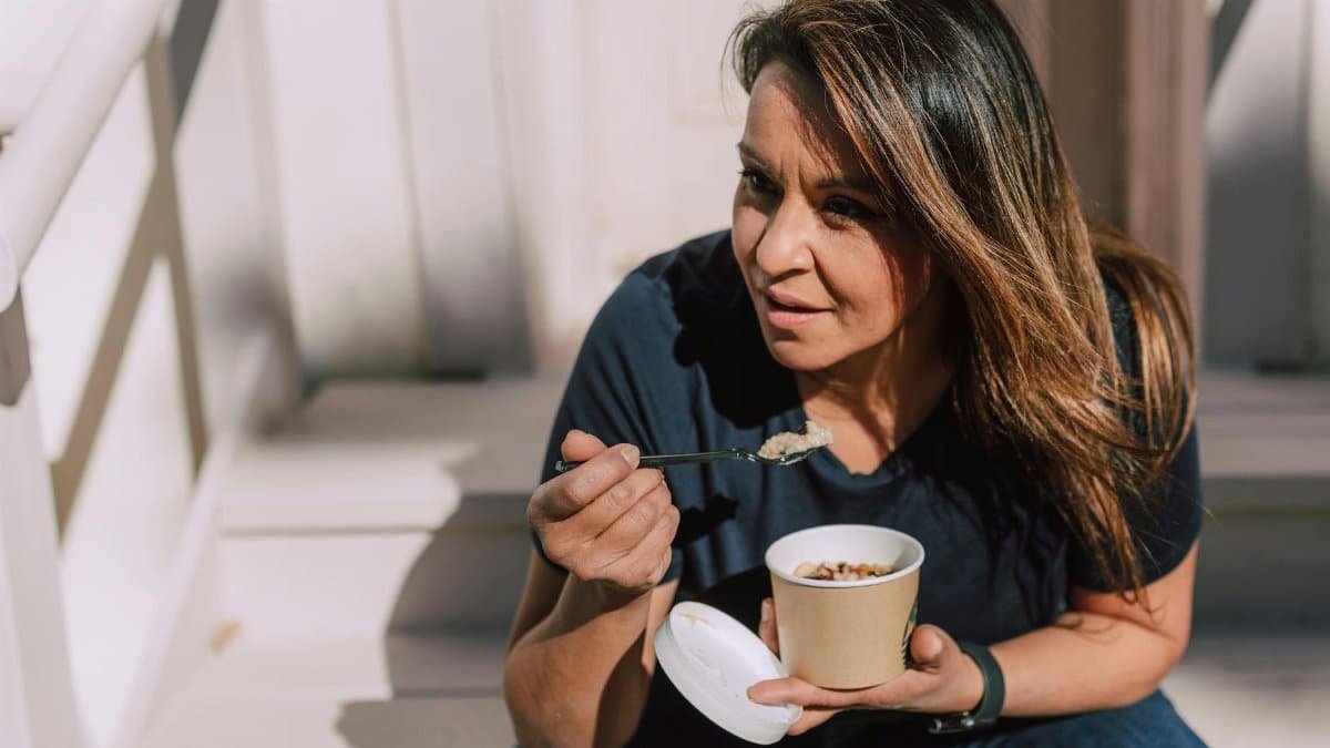 A woman in casual wear eating oats from a disposable bowl while sitting outdoors. Healthy lifestyle concept.
