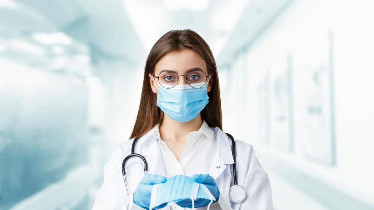 Young female doctor holding a face mask in hospital wearing gloves and stethoscope.
