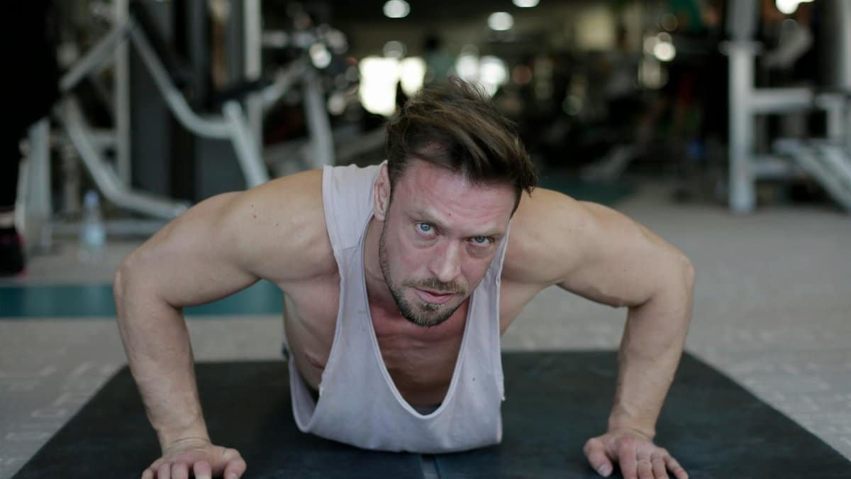 Determined adult muscular male athlete wearing sportswear performing handstand push up exercise during fitness workout on floor of modern spacious sport club and looking at camera