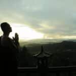 A monk in silhouette prays peacefully at sunrise with mountains in the background.