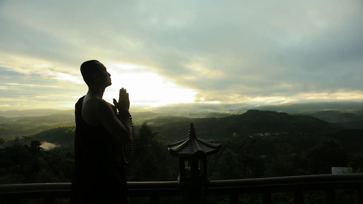 A monk in silhouette prays peacefully at sunrise with mountains in the background.