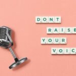 Studio shot of a vintage microphone and Scrabble tiles spelling 'Don't Raise Your Voice' on a pink background.