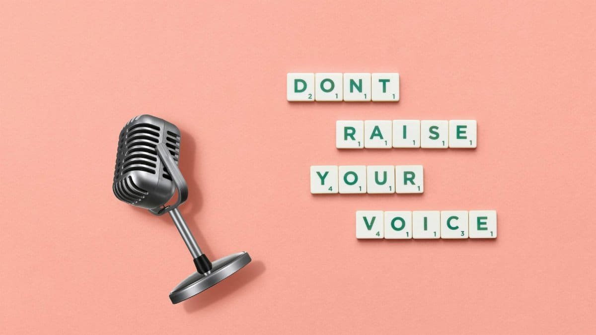 Studio shot of a vintage microphone and Scrabble tiles spelling 'Don't Raise Your Voice' on a pink background.
