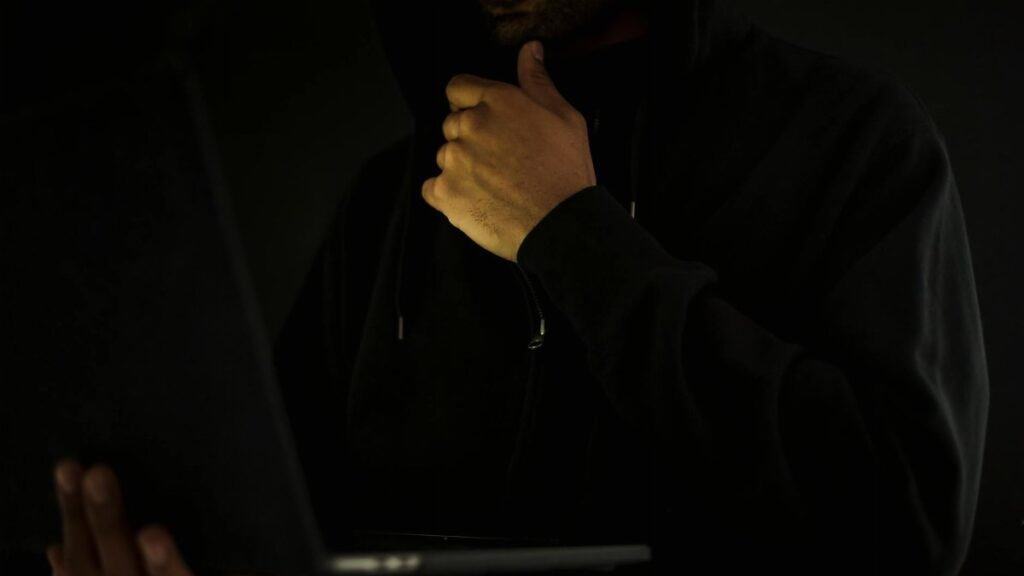 A man in a hoodie uses a laptop in darkness, symbolizing cybercrime and anonymity.
