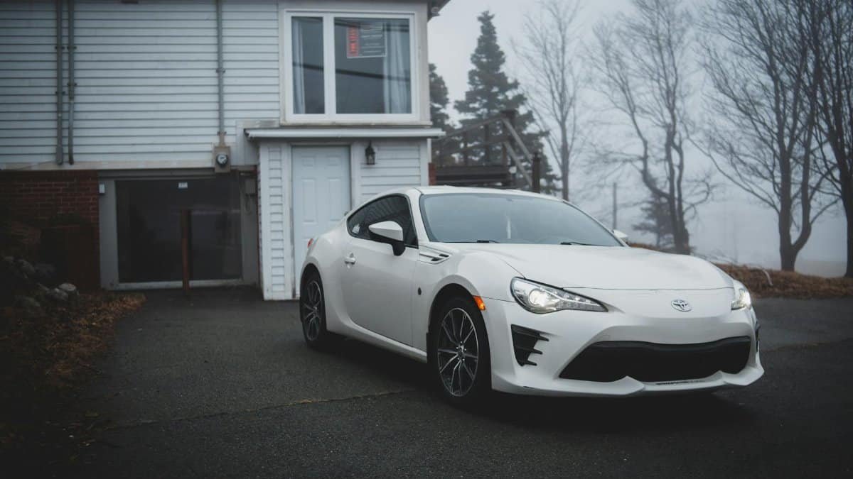 White sports car parked on driveway beside a modern house with foggy forest background.