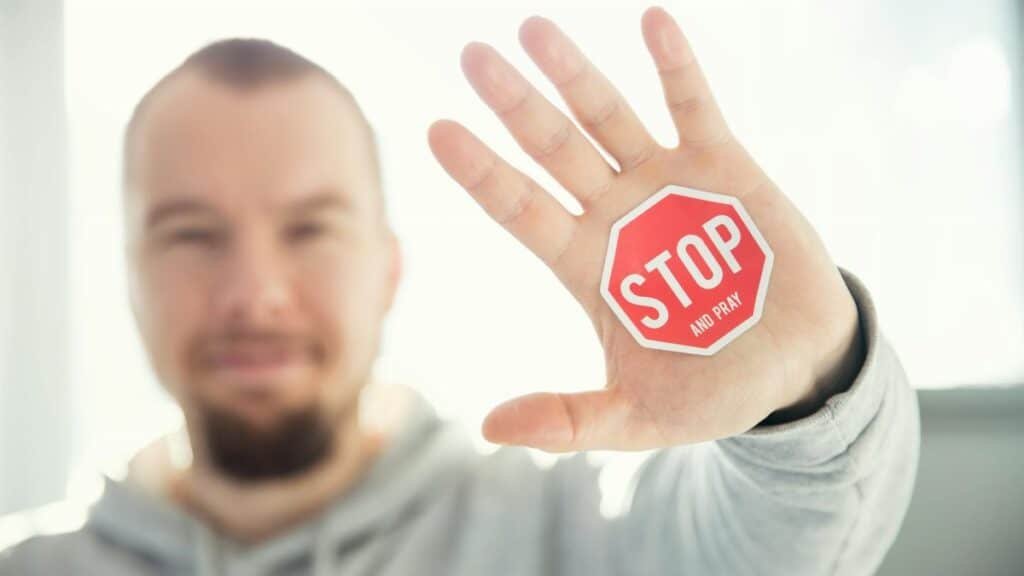 A man in a hoodie holds up his hand with a 'Stop and Pray' sticker. Focus on hand and sticker.