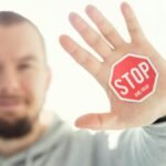 A man in a hoodie holds up his hand with a 'Stop and Pray' sticker. Focus on hand and sticker.