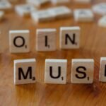 Scrabble tiles forming the words 'COIN' and 'MUSK' on a wooden table surface.