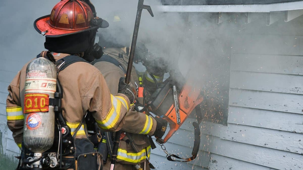 Firefighters using a power tool to cut through smoke-filled building exterior.