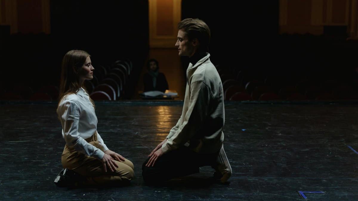 Two actors kneeling face-to-face in an emotional theater rehearsal, captured on a dimly lit stage.