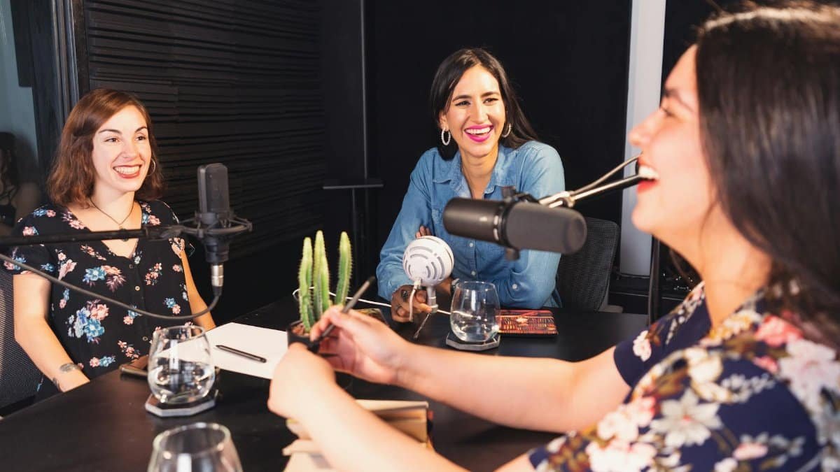 Three women sharing smiles and conversations during a podcast recording in a cozy studio setting.