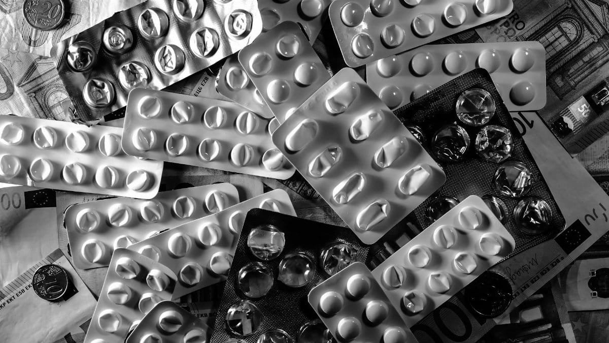Black and white image of pill blister packs scattered on euro bills and coins.