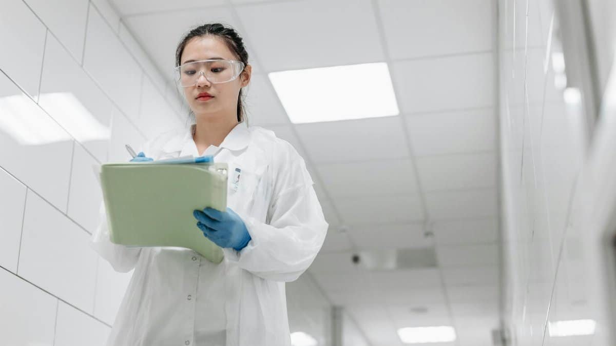 A focused female scientist in a lab coat writes notes in a laboratory hallway, showcasing professionalism in research settings.