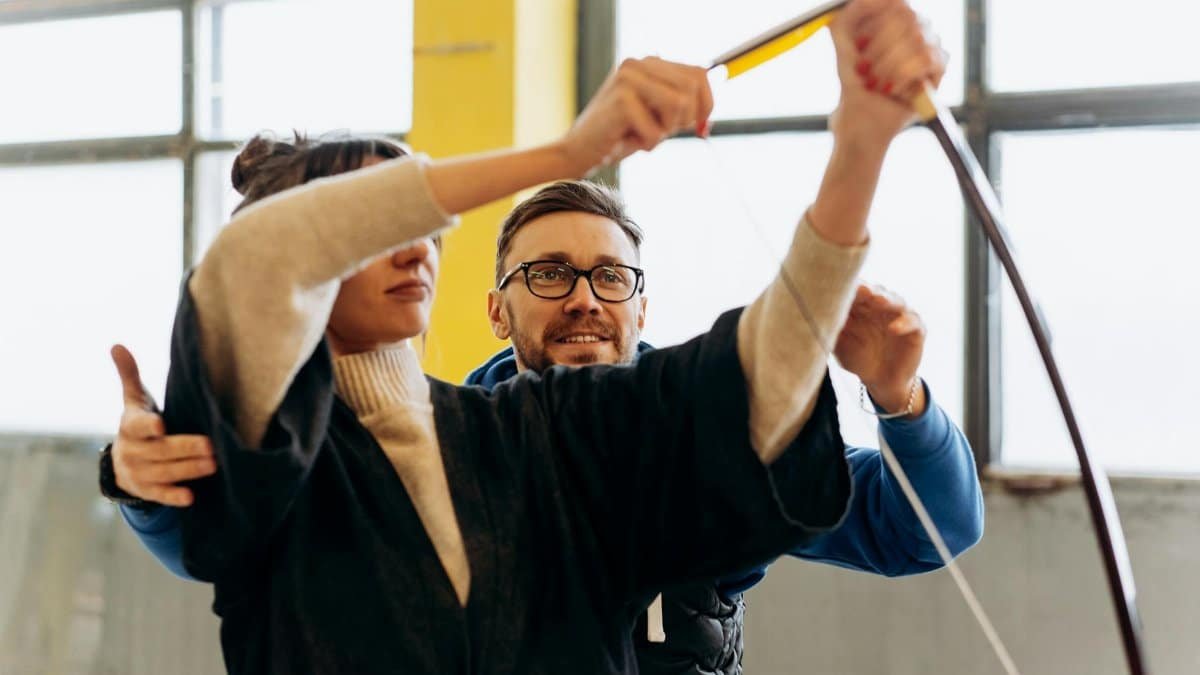 Adult woman learning archery with guidance from male coach indoors.