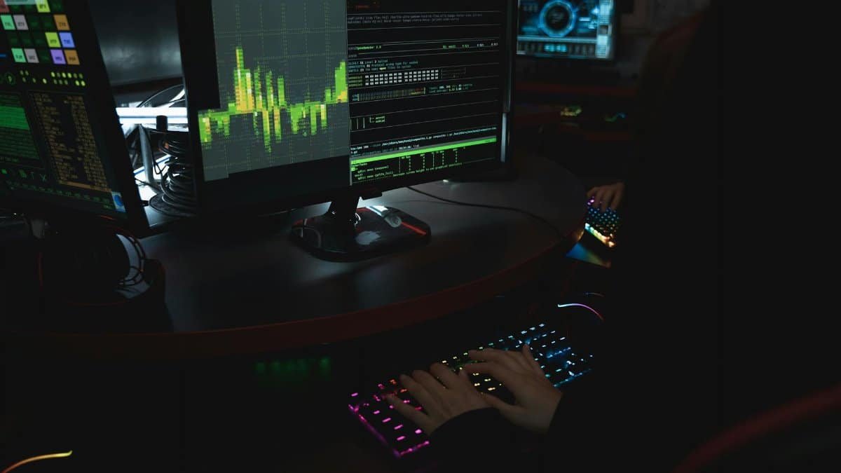 A cybersecurity expert in a dimly lit room is typing on a colorful keyboard with multiple screens displaying data.