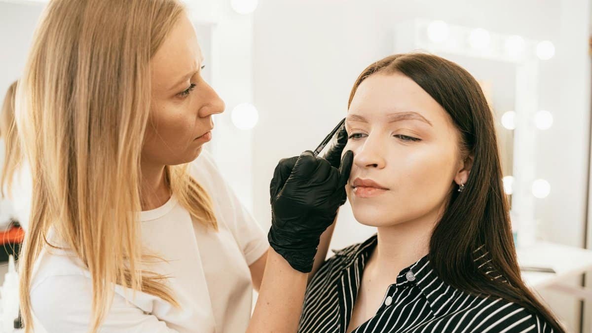 A skilled makeup artist applies eyebrow makeup on a client in a light-filled studio.