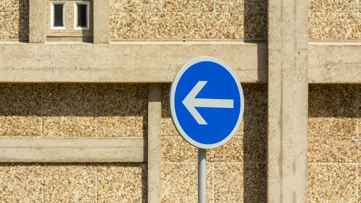 A blue traffic sign indicating a mandatory left turn in front of a textured urban wall.