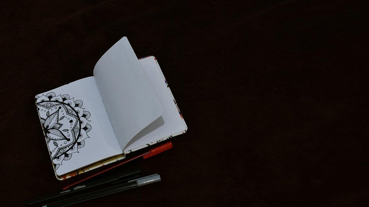 An open sketchbook with detailed doodle art next to pens on a dark surface, offering artistic inspiration.