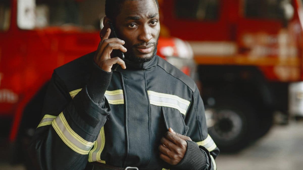 Close-up of a firefighter in protective gear making a phone call by a fire truck.