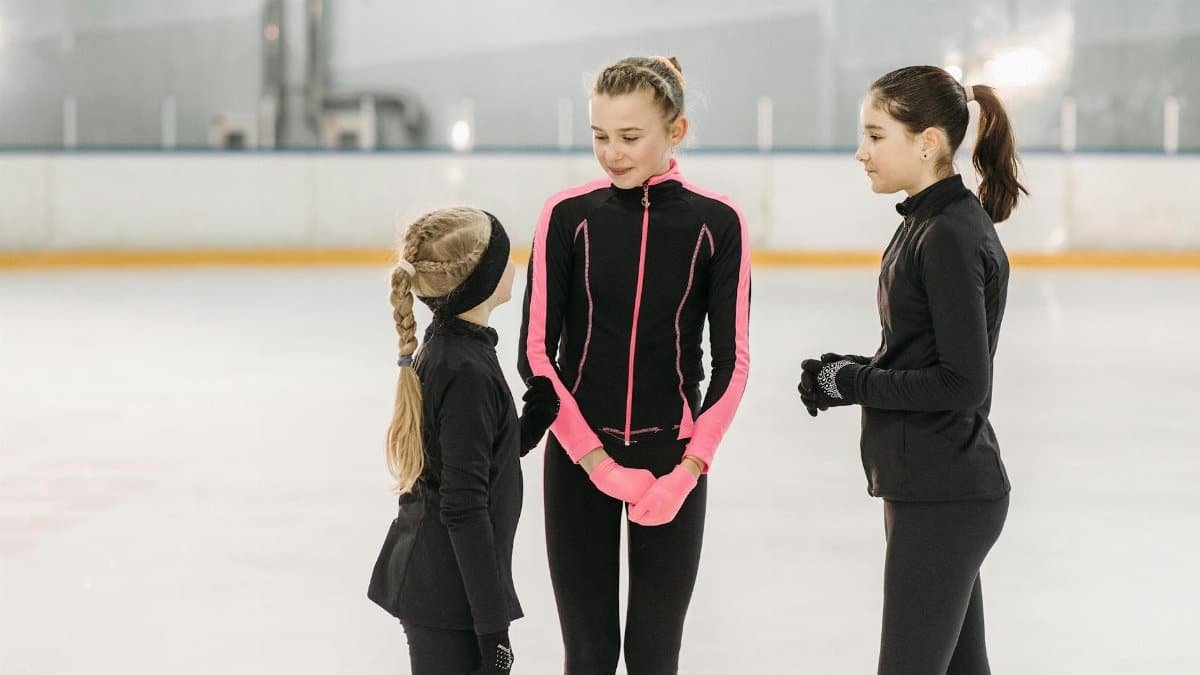 Three young female figure skaters in conversation on an indoor ice rink, dressed in black and pink outfits.