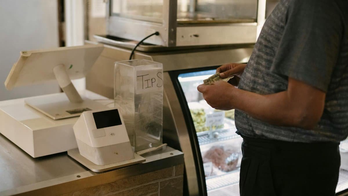 Unrecognizable male in casual clothes standing with money near cash register in grocery store while making purchase near glass showcase