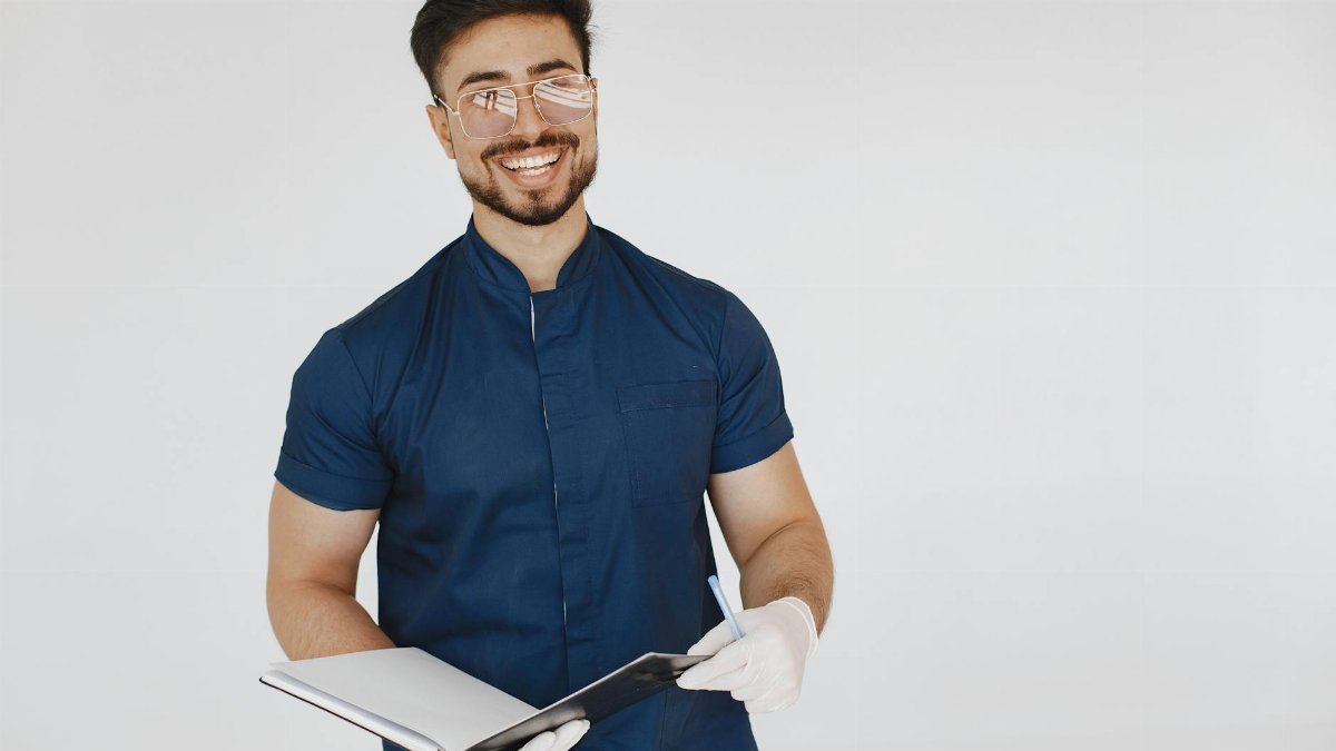 A cheerful medical professional in blue scrubs holding a notebook, wearing glasses and white gloves.