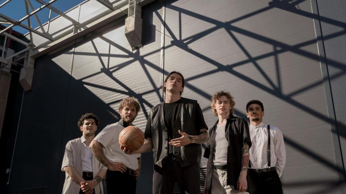 Five stylish men pose outside with a basketball, shadows creating a dynamic city backdrop.