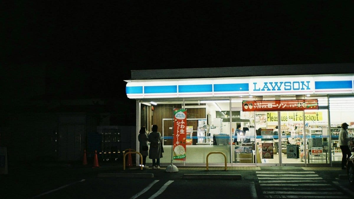 A nighttime view of a Lawson convenience store entrance with people.