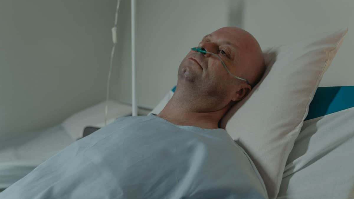 A bald man lying in a hospital bed wearing a nasal cannula for oxygen support, depicting illness and recovery.