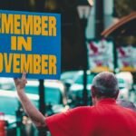 Man holding 'Remember in November' sign on urban street during the day.