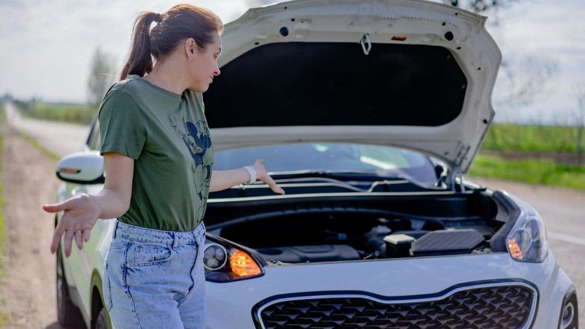 A woman looks puzzled by her broken car with an open hood on a rural road.