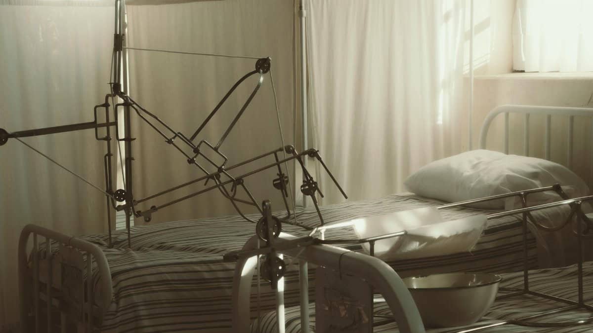 An empty hospital bed surrounded by medical equipment in a quiet, sunlit room.