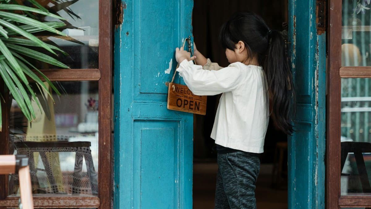 A young girl places an open sign on a rustic wooden door, emphasizing innocence and business.