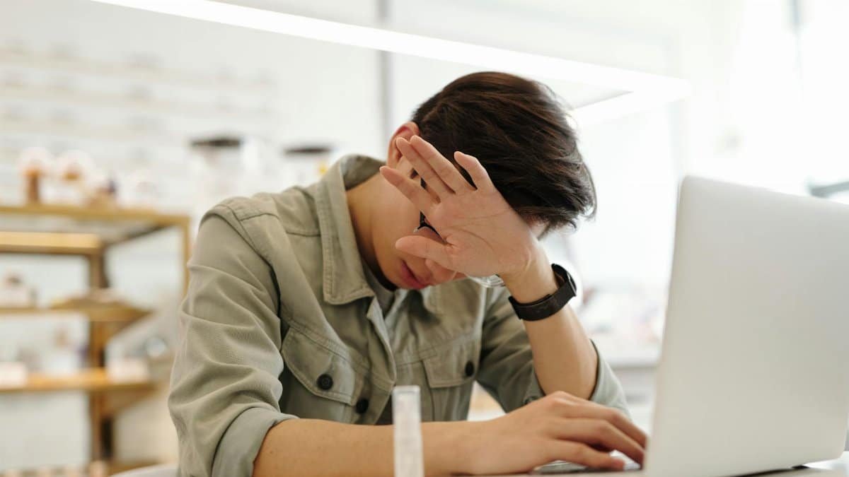 An adult man appears stressed while working on a laptop indoors, covering his face with one hand.