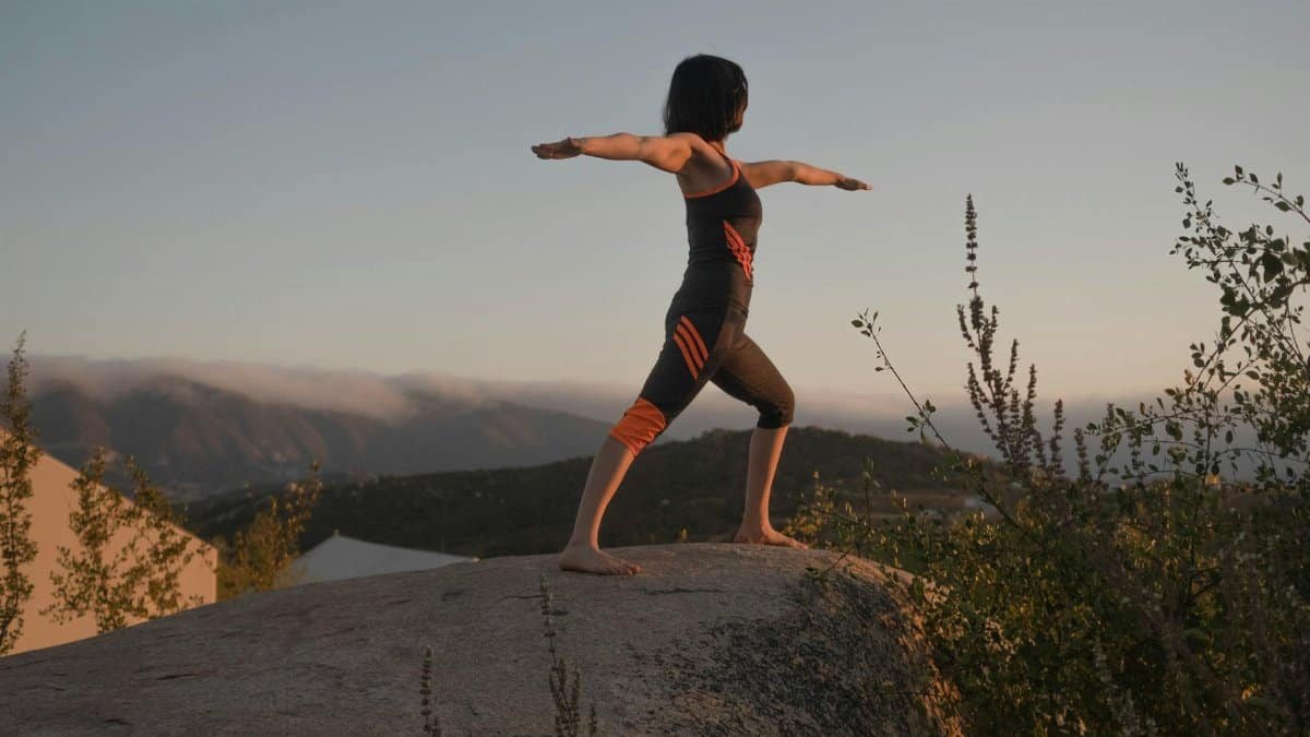 Woman in warrior II yoga pose on a rock at sunset, embracing wellness and mindfulness.