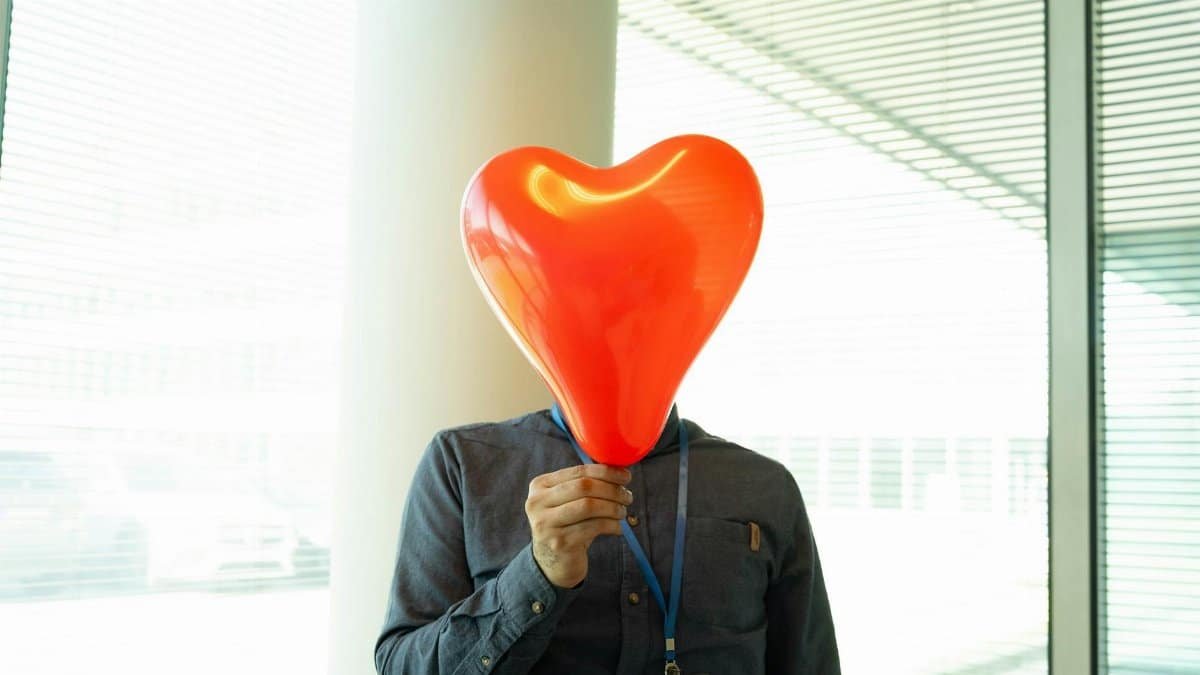 Humorous concept of a man holding a heart-shaped balloon over his face in an office setting.