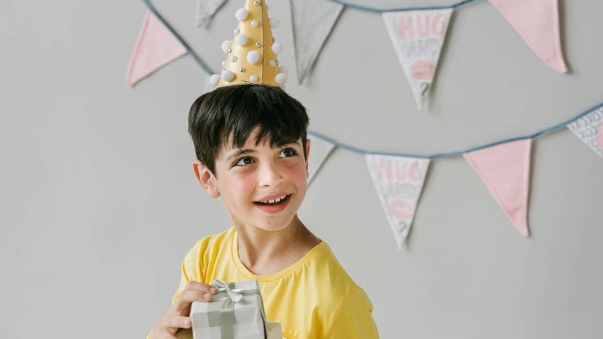 Young boy wearing party hat and holding a gift, celebrating birthday indoors.